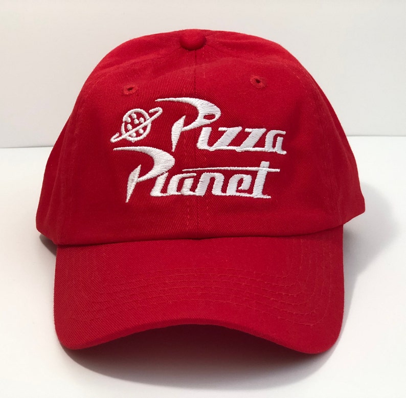 May include: A red baseball cap with white embroidered "Pizza Planet" text. The cap has a curved peak and a small button on top. The design includes a planet and a ring, a classic cap style.