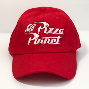 May include: A red baseball cap featuring the embroidered text "Pizza Planet" in white. The design includes a planet and a ring. The cap is made of a soft material and has a curved peak.
