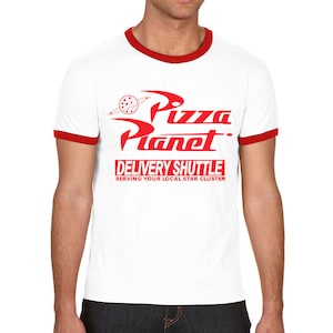 Puede incluir: Camiseta blanca con un ribete rojo y un gr&aacute;fico rojo del logotipo de Pizza Planet. El texto en el gr&aacute;fico dice "Pizza Planet Delivery Shuttle Serving Your Local Star Cluster".