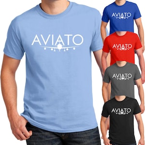 May include: A light blue t-shirt with the word "AVIATO" printed in white on the front. The word is centered and above a silhouette of an airplane. The t-shirt is available in a variety of colors.