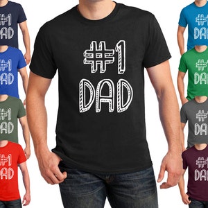 Op de afbeelding: Een zwart T-shirt met witte tekst die "#1 DAD" luidt in een gestileerd lettertype. Het shirt wordt getoond in tien verschillende kleuren: marineblauw, koninklijkblauw, legergroen, rood, saffier, Iers, houtskool, donkerrood en twee tinten groen.