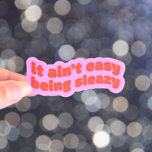 Könnte beinhalten: Ein pinkfarbener Aufkleber mit dem Schriftzug "it ain't easy being sleazy" in orangefarbener Schrift. Der Aufkleber wird vor einem verschwommenen Hintergrund aus weißen und blauen Bokeh-Lichtern gehalten.
