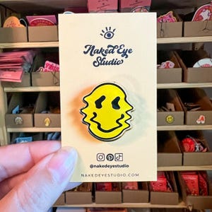 Könnte beinhalten: Gelber Smiley-Pin mit einem wellenförmigen, verzerrten Design. Der Pin befindet sich auf einer weißen Karte mit dem Text "Naked Eye Studio" und Social-Media-Handles.