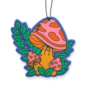 May include: A colorful air freshener with a mushroom design. The air freshener features a large pink mushroom with orange spots and a yellow stem, surrounded by green leaves and a blue border. A black string is attached for hanging.