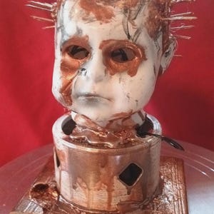 May include: A doll head sculpture with a distressed appearance. The white face has bronze-colored accents around the eyes and mouth, with metal spikes protruding from the head. The sculpture sits on a bronze-colored cylindrical base.