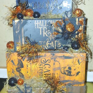 May include: A stack of decorative Halloween-themed boxes. The boxes are painted in black, orange, and blue with text and spooky embellishments like spiders, skulls, and glittery orbs. The top box has a skull on top.
