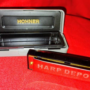 May include: A black and red harmonica with the text "HARP DEPOT" and a gray case with the name "HOHNER" in gold lettering. The harmonica is resting on a red surface.