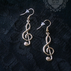 Könnte beinhalten: Ein Paar goldfarbene Ohrringe mit einem Violinschlüssel-Design. Die Ohrringe befinden sich auf einem schwarzen Hintergrund.