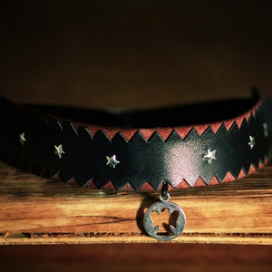 May include: A black leather choker with a red leather trim and silver star accents. The choker has a silver pendant with a star-shaped cutout.