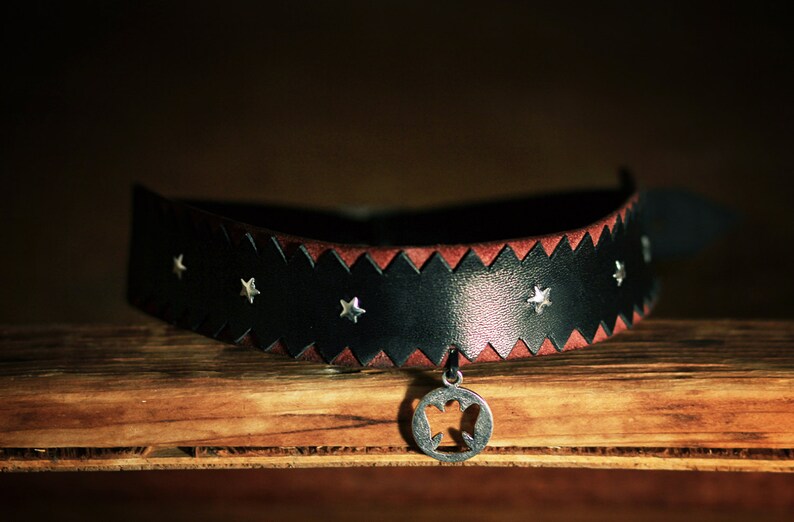 May include: A black leather choker with a red leather trim and silver star accents. The choker has a silver pendant with a star-shaped cutout.