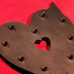 May include: A black leather heart-shaped paddle with metal studs and a heart-shaped cutout in the center. The paddle is on a red background.