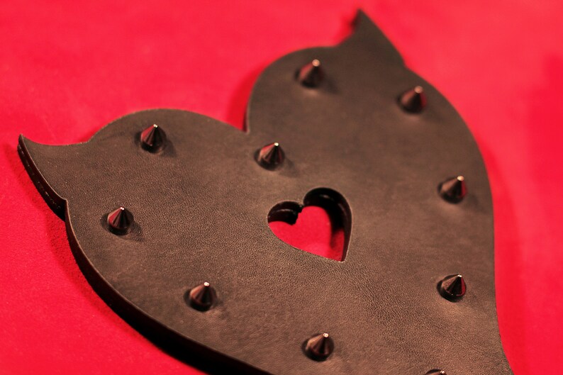 May include: A black leather heart-shaped paddle with metal studs and a heart-shaped cutout in the center. The paddle is on a red background.