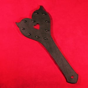 May include: A black leather paddle shaped like a heart with a smaller heart cut out in the center. The paddle is studded with metal rivets.