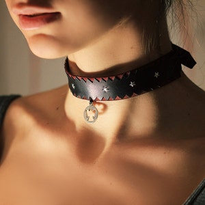May include: A black leather choker with a silver star design and a silver pendant. The choker is worn by a person with a light complexion.