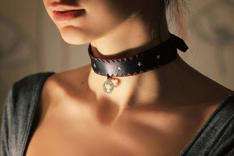 May include: A black leather choker with a silver star design and a silver pendant. The choker is worn by a person with a light complexion.