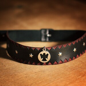 May include: A black leather choker with a red stitching detail and silver star accents. A silver pendant with an angel cutout hangs from the center.