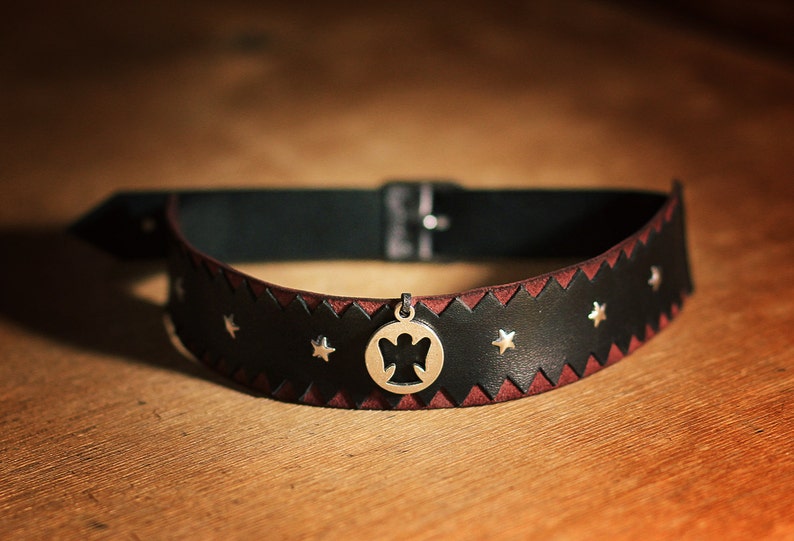 May include: A black leather choker with a red stitching detail and silver star accents. A silver pendant with an angel cutout hangs from the center.