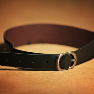 May include: A black leather belt with a silver buckle. The belt is lying on a brown surface.