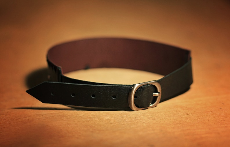 May include: A black leather belt with a silver buckle. The belt is lying on a brown surface.