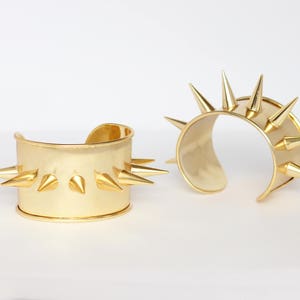 Może przedstawiać: Dwa złote metalowe bransoletki z ostrymi kolcami. Bransoletki są otwarte i mają szeroki, płaski pasek.