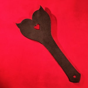 May include: A brown leather paddle shaped like a heart with a smaller heart cut out in the center. The paddle is on a red background.