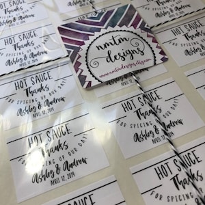 May include: White square stickers with black text that reads "Hot Sauce Thanks for Spicing Up Our Day! Ashley & Andrew April 12, 2019". The stickers are arranged in rows on a white surface. A purple and white watercolor design with the text "nmtm designs www.nmtmdesigns.etsy.com Ashley & Andrew April 12, 2019" is in the center of the image.