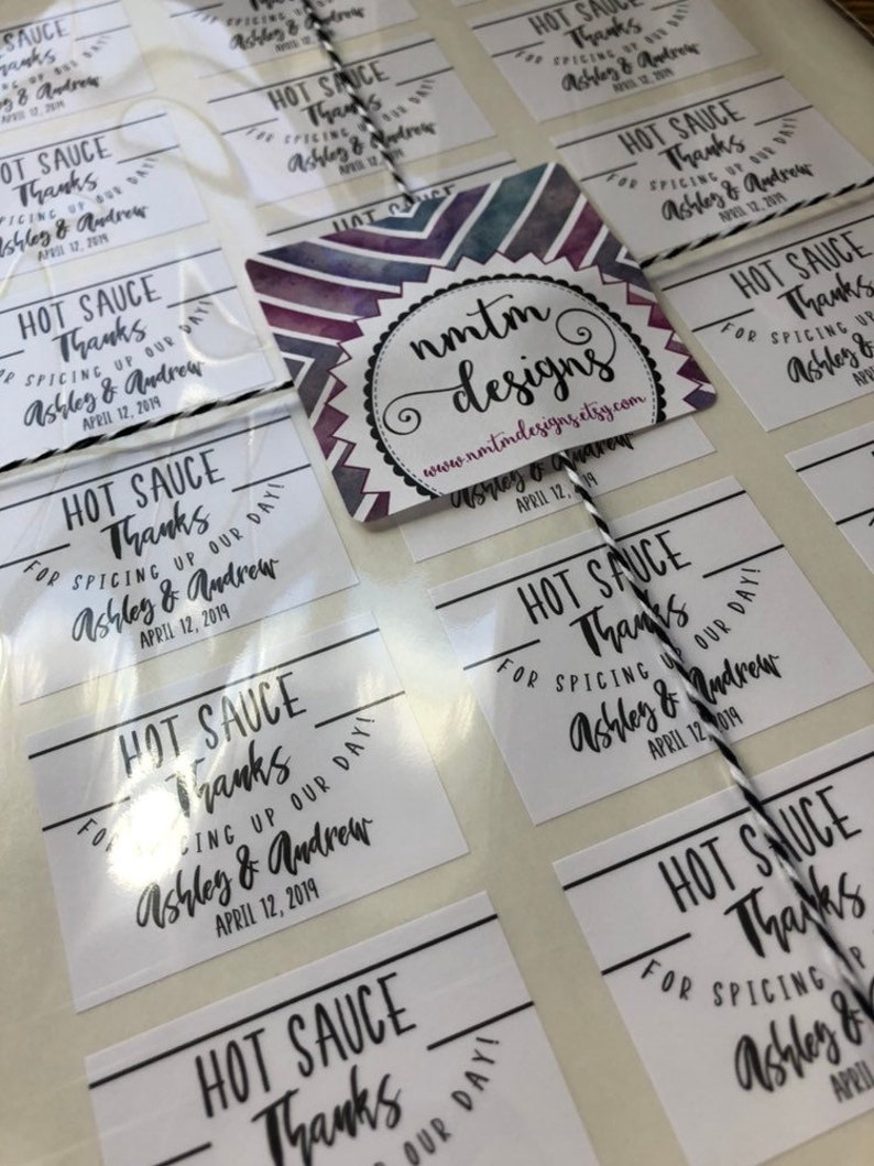 May include: White square stickers with black text that reads "Hot Sauce Thanks for Spicing Up Our Day! Ashley & Andrew April 12, 2019". The stickers are arranged in rows on a white surface. A purple and white watercolor design with the text "nmtm designs www.nmtmdesigns.etsy.com Ashley & Andrew April 12, 2019" is in the center of the image.