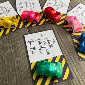 May include: A set of five cards with a yellow and black striped background. Each card has a different colored toy car attached to it. The cards have the text "I'm wheelic GLAD WE ARE friends! Love, Evie" on them.