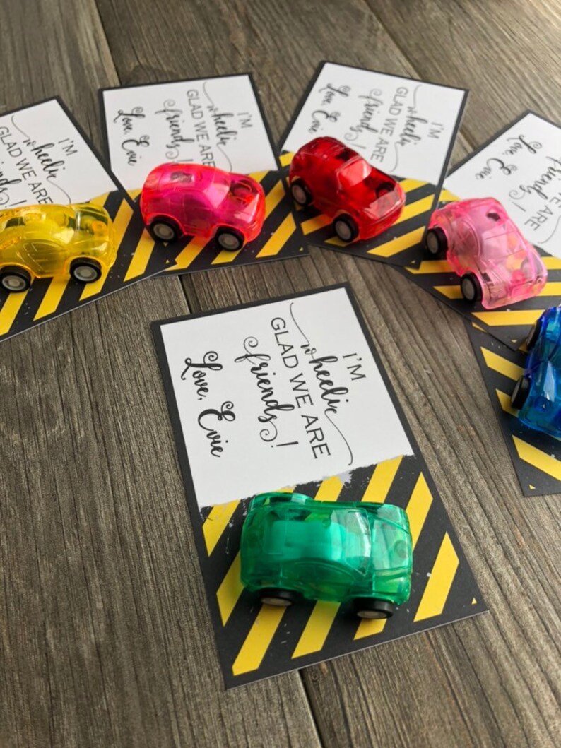 May include: A set of five cards with a yellow and black striped background. Each card has a different colored toy car attached to it. The cards have the text "I'm wheelic GLAD WE ARE friends! Love, Evie" on them.