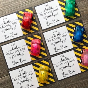 May include: Five colorful toy cars with wheels are attached to white cards with black borders. The cards have black and yellow striped borders. The cards have the text "I'm wheelic GLAD WE ARE friends! Love, Evie" printed on them.