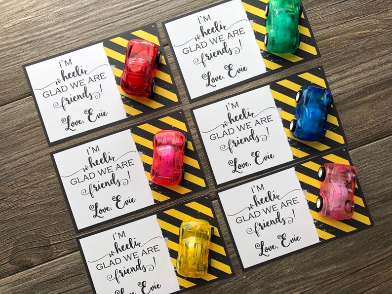 May include: Five colorful toy cars with wheels are attached to white cards with black borders. The cards have black and yellow striped borders. The cards have the text "I'm wheelic GLAD WE ARE friends! Love, Evie" printed on them.