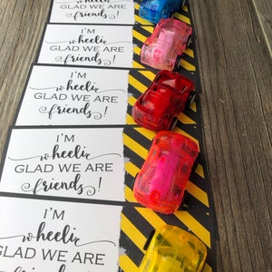 May include: Six small, clear plastic toy cars in different colors, lined up on a black and yellow striped background. Each car has a white card attached to it with the text "I'm wheelie glad we are friends!"