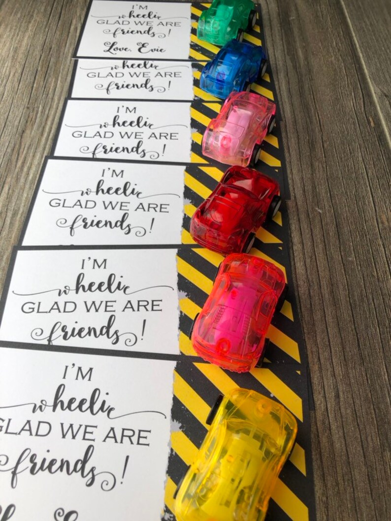 May include: Six small, clear plastic toy cars in different colors, lined up on a black and yellow striped background. Each car has a white card attached to it with the text "I'm wheelie glad we are friends!"