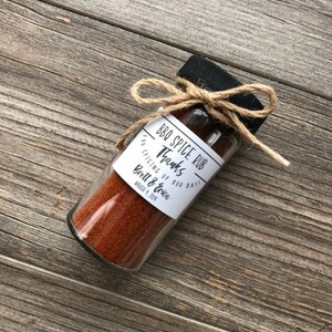 May include: A small glass jar filled with red BBQ spice rub, tied with twine and a label that reads "BBQ Spice Rub Thanks for spicing up our day! Brett & Erica March 9, 2019".
