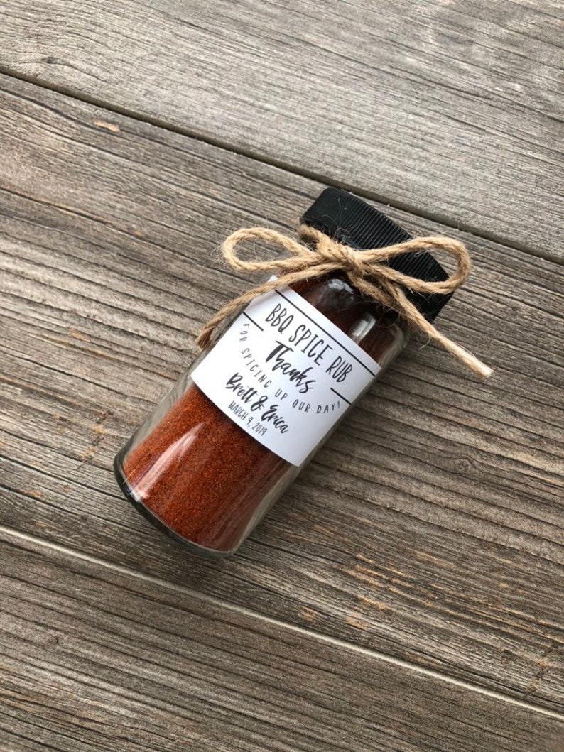 May include: A small glass jar filled with red BBQ spice rub, tied with twine and a label that reads "BBQ Spice Rub Thanks for spicing up our day! Brett & Erica March 9, 2019".