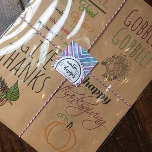 Op de afbeelding: Een set van vier bruine kraftpapierkaarten met Thanksgiving-thema illustraties en tekst. De kaarten bevatten een kalkoen, een pompoen, herfstbladeren en de woorden "Give Thanks", "Happy Thanksgiving", "Drink & Be Thankful", "Gobble Gobble" en "Great Food Wines Friends".