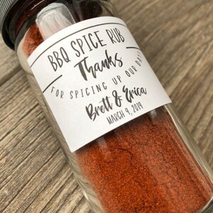 May include: A glass jar filled with red BBQ spice rub with a white label that reads "BBQ Spice Rub Thanks for spicing up our day! Brett & Erica March 9, 2019"