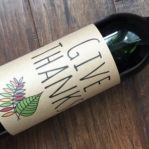 Op de afbeelding: Een bruine wijnfles met een papieren etiket waarop "GIVE THANKS" staat met een kleurrijk bladontwerp.