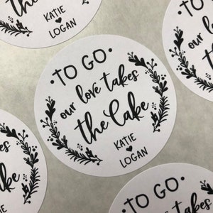 May include: White round stickers with black text and floral designs. The text reads "To Go. Our love takes the Cake" with the names Katie and Logan.