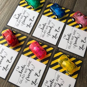 May include: A set of six small, colorful toy cars with a white card attached to each. The cards have black text that reads "I'm wheelic GLAD WE ARE friends! Love, Evie". The cards are on a yellow and black striped background.
