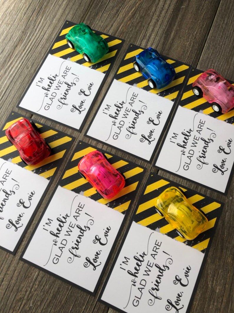 May include: A set of six small, colorful toy cars with a white card attached to each. The cards have black text that reads "I'm wheelic GLAD WE ARE friends! Love, Evie". The cards are on a yellow and black striped background.