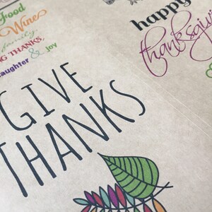 Op de afbeelding: Een bruine papieren achtergrond met zwarte tekst die "Give Thanks" luidt met een kleurrijk bladontwerp. De tekst "Happy Thanksgiving" is in paarse inkt geschreven.