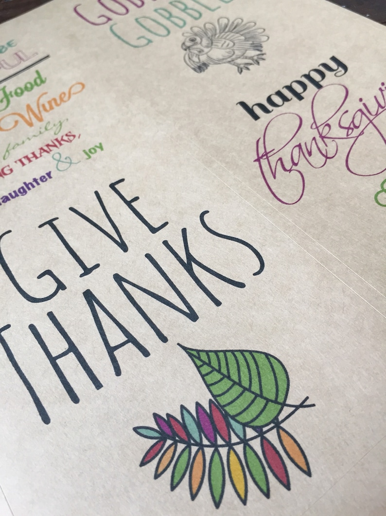 Op de afbeelding: Een bruine papieren achtergrond met zwarte tekst die "Give Thanks" luidt met een kleurrijk bladontwerp. De tekst "Happy Thanksgiving" is in paarse inkt geschreven.