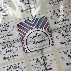 May include: A set of white square stickers with black text that reads "Hot Sauce Thanks for Spicing Up Our Day! Ashley & Andrew April 12, 2019". The stickers are arranged in rows and columns on a white surface. A larger sticker with a watercolor design and the text "nmtm designs www.nmtmdesigns.etsy.com April 12, 2019" is in the center of the image.