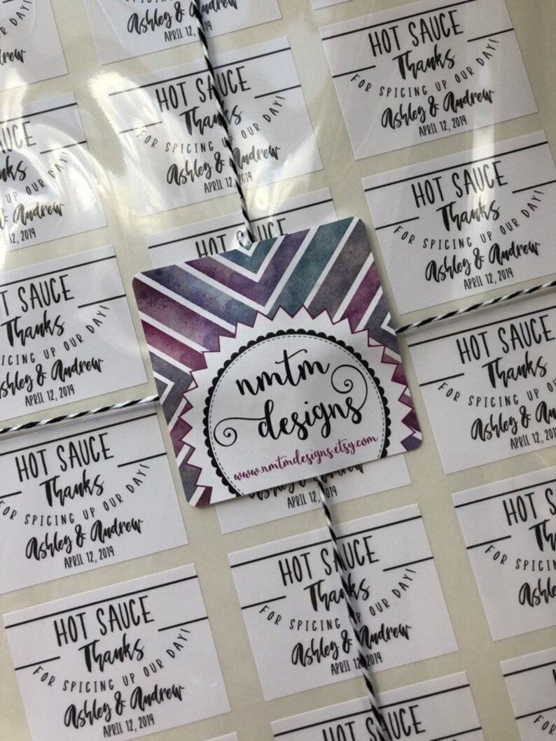 May include: A set of white square stickers with black text that reads "Hot Sauce Thanks for Spicing Up Our Day! Ashley & Andrew April 12, 2019". The stickers are arranged in rows and columns on a white surface. A larger sticker with a watercolor design and the text "nmtm designs www.nmtmdesigns.etsy.com April 12, 2019" is in the center of the image.