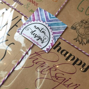 Op de afbeelding: Een bruin papieren pakketje dat vastgebonden is met touw en een paars lint. Het pakketje heeft een sticker met een aquarelontwerp en de tekst "nmtm designs" en "www.nmtmdesigns.etsy.com". Het pakketje heeft ook de tekst "happy thanksgiving" erop gedrukt.