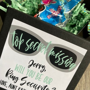 May include: A black box with a white card inside. The card has a green and black illustration of sunglasses with the text "Top Secret Mission" and "Jerry, Will You Be Our Ring Security?"  Love, Aunt Kathy & Uncle Matt.