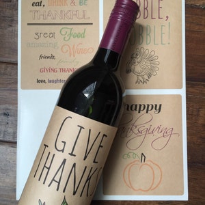 Op de afbeelding: Een bruin papieren wijnfleslabel met de tekst "Give Thanks" en een kleurrijk bladerenontwerp. Het label is bevestigd aan een fles rode wijn.
