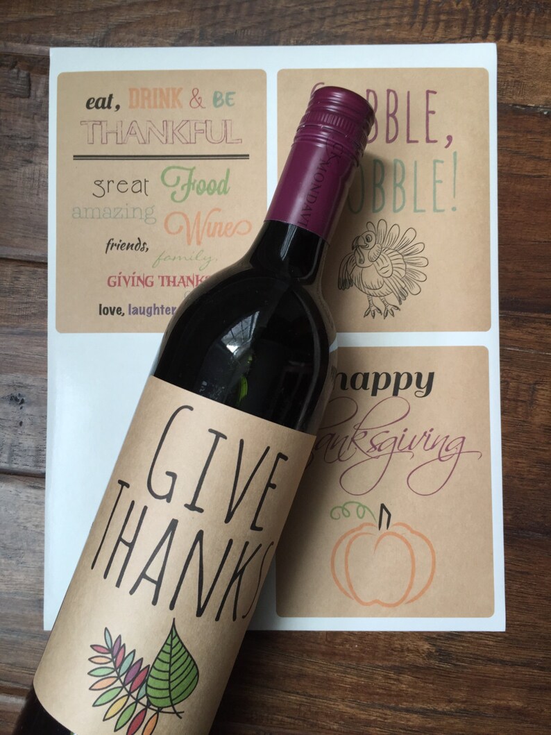 Op de afbeelding: Een bruin papieren wijnfleslabel met de tekst "Give Thanks" en een kleurrijk bladerenontwerp. Het label is bevestigd aan een fles rode wijn.