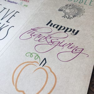 Op de afbeelding: Een bruine papieren kaart met de woorden "GOBBLE GOBBLE" in groene tekst. De kaart heeft ook een zwart-witte illustratie van een kalkoen. Onder de kalkoen staat de tekst "happy thanksgiving" in paarse en zwarte tekst. Een kleine oranje pompoen met een groene steel bevindt zich onderaan de kaart.
